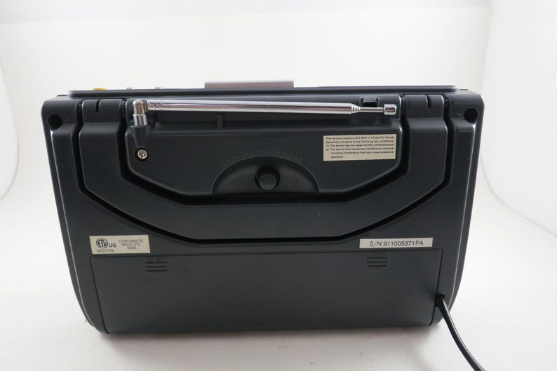 May include: A black portable radio with a silver antenna extended. The back of the radio features a control knob, a small information label, and a power cord. The radio is set against a white background.