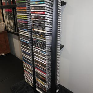May include: A tall, black, vertical media storage rack holding two columns of stacked CD jewel cases. The rack features a slim, minimalist frame design with a central support bar, displaying a large collection of music albums with various colored spines.
