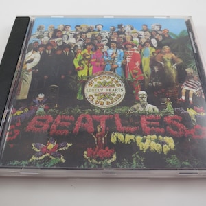 May include: A CD case featuring the album cover of The Beatles' "Sgt. Pepper's Lonely Hearts Club Band." The cover art displays a crowd of figures, the band members, and the album title spelled out in flowers.