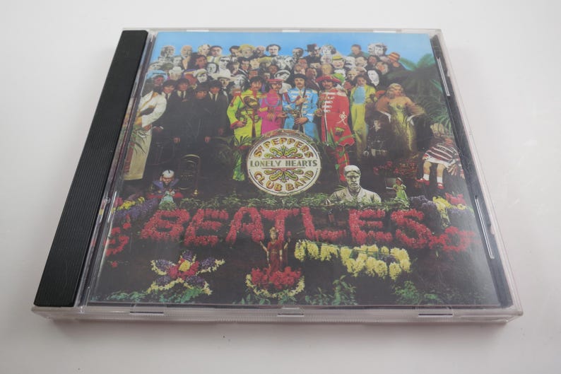 May include: A CD case featuring the album cover of The Beatles' "Sgt. Pepper's Lonely Hearts Club Band." The cover art displays a crowd of figures, the band members, and the album title spelled out in flowers.