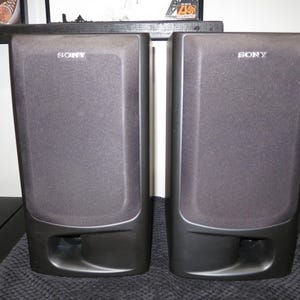 May include: A pair of black Sony bookshelf speakers with rectangular fabric grilles covering the drivers and a prominent bass reflex port located at the bottom of each unit. The Sony brand name is printed in white at the top of each speaker.