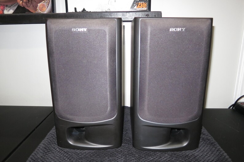 May include: A pair of black Sony bookshelf speakers with rectangular fabric grilles covering the drivers and a prominent bass reflex port located at the bottom of each unit. The Sony brand name is printed in white at the top of each speaker.