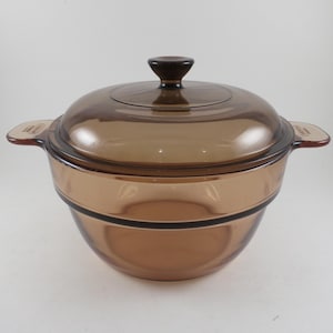 May include: A round, amber-colored glass casserole dish with a matching lid. The dish features two integrated side handles and a central knob on the lid. The glass is translucent and brown, with a smooth, curved design and a horizontal band detail.