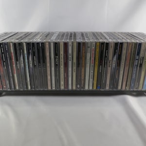 May include: A long, rectangular black wooden rack holding a large collection of music CDs in clear plastic jewel cases. The spines of the CDs are visible, displaying various titles, artist names, and graphic designs in a wide array of colors.