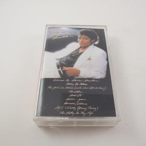 May include: A vintage cassette tape featuring Michael Jackson's album "Thriller." The cover shows a photo of Michael Jackson in a white jacket. The tracklist includes "Wanna Be Startin' Somethin'" and "Thriller."