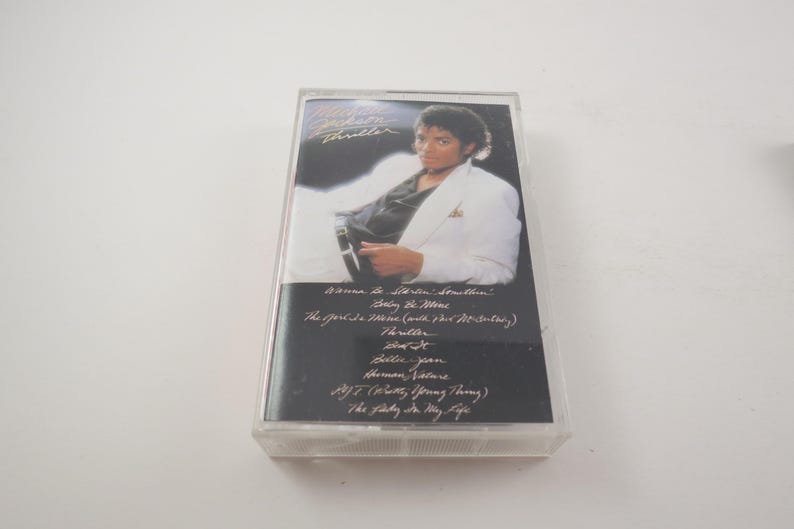 May include: A vintage cassette tape featuring Michael Jackson's album "Thriller." The cover shows a photo of Michael Jackson in a white jacket. The tracklist includes "Wanna Be Startin' Somethin'" and "Thriller."