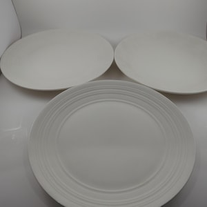 May include: Four white ceramic plates. Three are oval-shaped, and one is round with concentric ring detailing. The plates are arranged on a white surface, suggesting a dining or kitchen setting. The plates are clean and simple.