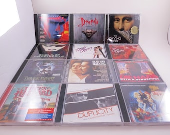 May include: A collection of twelve movie soundtrack CDs in clear plastic jewel cases arranged in a grid. Titles include Days of Thunder, Dracula, The Da Vinci Code, Dead Presidents, Dirty Dancing, Demon Knight, Dead Man Walking, Die Hard, The Dukes of Hazzard, Duplicity, and a James Bond film.