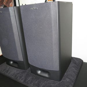 May include: A pair of black Sony speakers with a dark grey fabric front. The speakers are rectangular with rounded edges and a small opening at the bottom. The Sony logo is visible on the top of each speaker.
