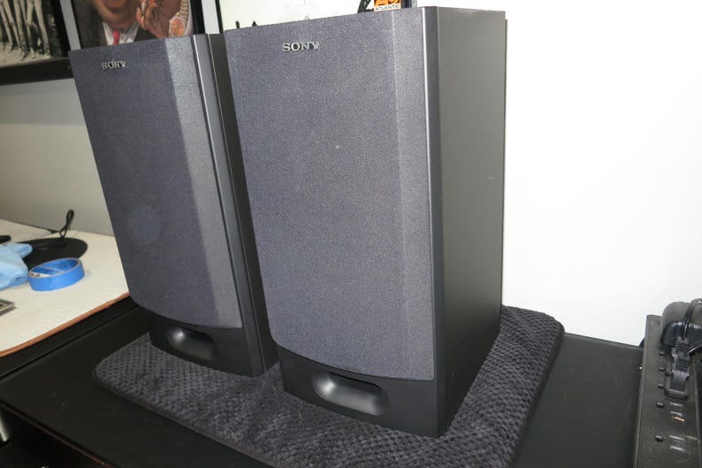 May include: A pair of black Sony speakers with a dark grey fabric front. The speakers are rectangular with rounded edges and a small opening at the bottom. The Sony logo is visible on the top of each speaker.