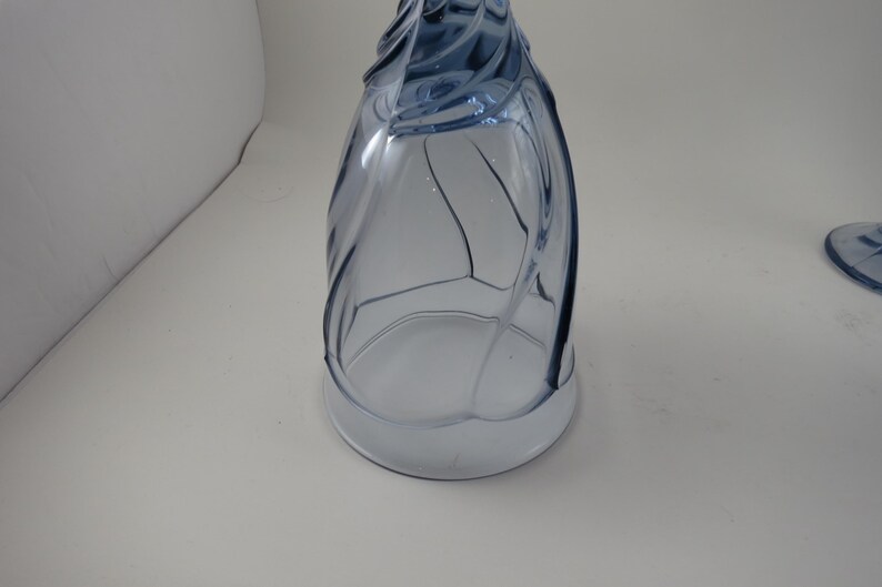 Pu&ograve; includere: Un bicchiere da vino blu in vetro con un design unico a spirale. Il bicchiere ha una base ampia e un gambo alto e curvo. Il bicchiere &egrave; in vetro trasparente con una sfumatura blu chiaro. Il bicchiere &egrave; vuoto e su una superficie bianca.