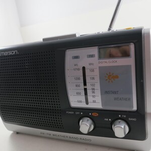 May include: A black and silver Emerson AM/FM weather band radio. The radio has a digital clock and instant weather display. The radio has a telescoping antenna and two silver knobs for power and band selection.