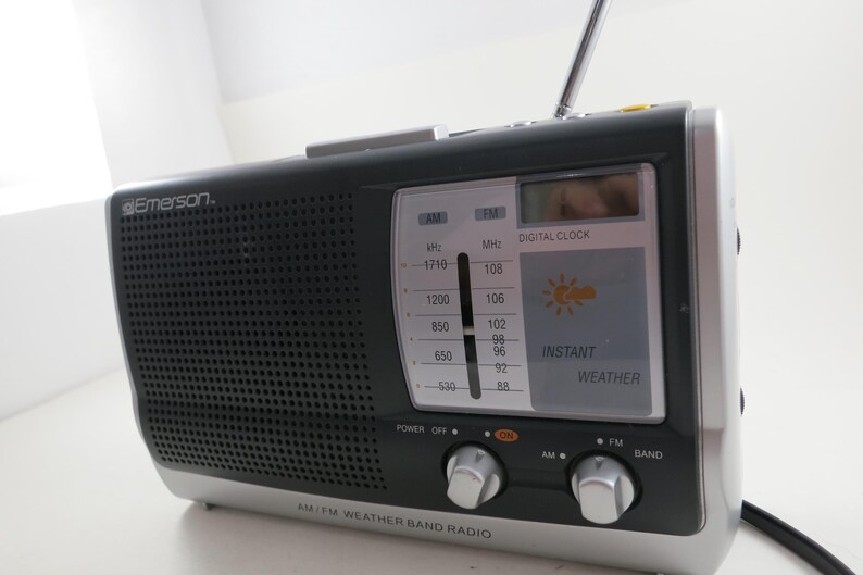 May include: A black and silver Emerson AM/FM weather band radio. The radio has a digital clock and instant weather display. The radio has a telescoping antenna and two silver knobs for power and band selection.