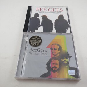 May include: Two Bee Gees music CDs in clear plastic cases. The top case features a white cover with black silhouettes of three figures. The bottom case is titled Number Ones and displays a colorful graphic of the band members with a circular sticker.
