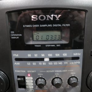 May include: Black Sony stereo system with a digital display showing the time. Features include FM/AM radio, CD player, and rotary equalizer. Buttons for play, pause, and tuning are visible.