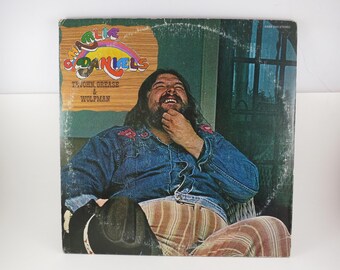 May include: A vinyl record cover featuring Charlie Daniels with the text Charlie Daniels, Te John, Grease & Wolfman. The image shows a bearded individual wearing a blue denim shirt with red floral embroidery and striped pants, holding a black hat.