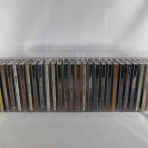 May include: A clear acrylic storage rack containing a long row of music CDs in their plastic jewel cases. The rack is rectangular and holds the discs upright, displaying the spines of various albums with assorted text and graphic designs.