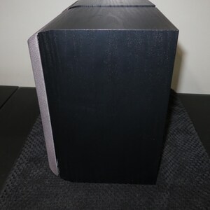 May include: A black speaker with a gray accent on the side. The speaker has a rectangular shape with a slightly angled top. The speaker is sitting on a dark surface.