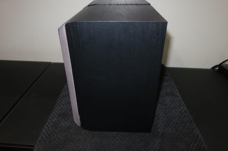 May include: A black speaker with a gray accent on the side. The speaker has a rectangular shape with a slightly angled top. The speaker is sitting on a dark surface.