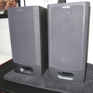May include: Two grey Sony speakers with a rectangular shape and a dark grey fabric covering the front. Each speaker has a rectangular opening at the bottom. The Sony logo is visible at the top of each speaker.