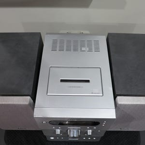 May include: A silver stereo system with two black speakers on either side. The stereo has a CD player and a control panel with knobs and buttons. The speakers are rectangular and sit on top of the stereo's base.