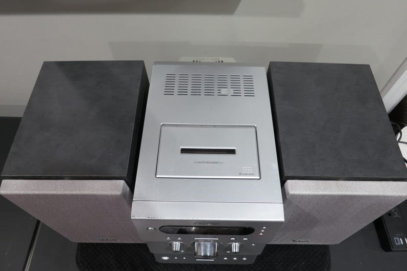 May include: A silver stereo system with two black speakers on either side. The stereo has a CD player and a control panel with knobs and buttons. The speakers are rectangular and sit on top of the stereo's base.