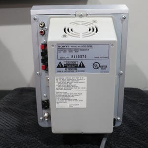 May include: Rear view of a silver and gray Sony compact disc deck receiver. The back panel features various input/output ports, a cooling fan, and a label with model and serial number information. The unit is on a black surface.