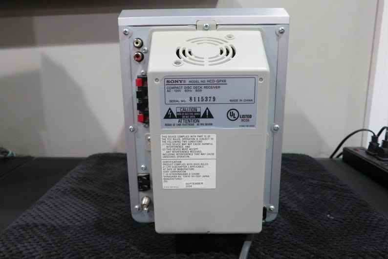 May include: Rear view of a silver and gray Sony compact disc deck receiver. The back panel features various input/output ports, a cooling fan, and a label with model and serial number information. The unit is on a black surface.