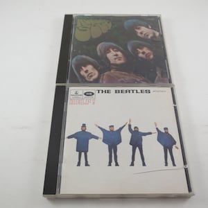 May include: Two compact disc cases. The top case features a photo of four men with long hair. The bottom case has the text "THE BEATLES" and "HELP!" with a photo of four men in blue coats.