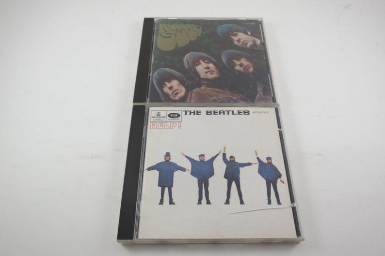 May include: Two compact disc cases. The top case features a photo of four men with long hair. The bottom case has the text "THE BEATLES" and "HELP!" with a photo of four men in blue coats.