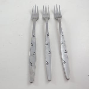 May include: Three silver forks with a floral design on the handles. The forks are arranged on a white surface. Each fork has four tines and a long, slender handle with a repeating pattern of small floral motifs.