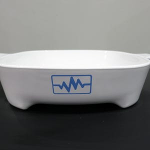 May include: White, oval-shaped baking dish with a blue graphic of a heartbeat. The dish has two handles and four small feet. The dish is made of a glossy material.