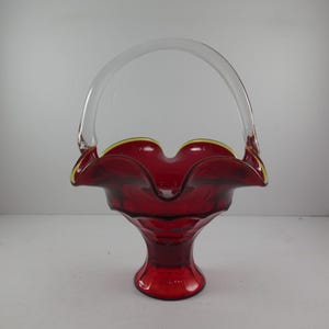 May include: A ruby red glass basket with a clear glass handle. The basket has a ruffled rim with a gold trim. The base is a pedestal shape. This decorative piece is suitable for home decor.