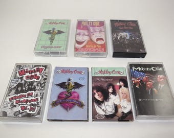 May include: A collection of seven Motley Crue cassette tapes. The tapes feature album art with various designs, including band photos, a heart with wings, and the American flag. The cassette cases are clear plastic.