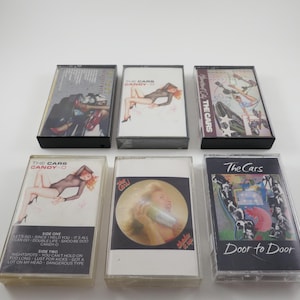 May include: Six cassette tapes by the band The Cars. The tapes are in clear plastic cases with black labels. The titles of the tapes are "Candy-O", "The Cars", "Door to Door", "Candy-O", "The Cars", and "The Cars".