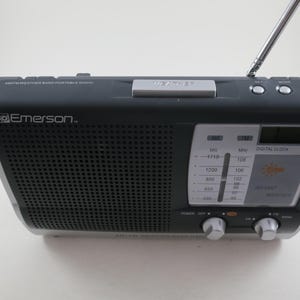May include: A black and silver Emerson AM/FM weather band portable radio. The radio has a digital clock, a telescoping antenna, and a tuning dial. The front of the radio has a speaker grill and a display with weather information.