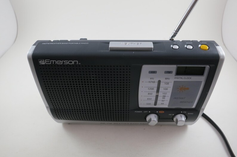 May include: A black and silver Emerson AM/FM weather band portable radio. The radio has a digital clock, a telescoping antenna, and a tuning dial. The front of the radio has a speaker grill and a display with weather information.