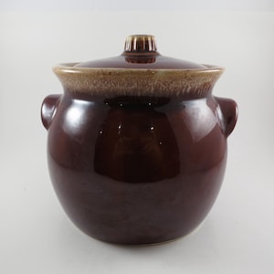 May include: A brown ceramic bean pot with a lid. The pot has two handles and a light brown rim. The lid has a small knob on top. The bean pot is a classic kitchenware item.