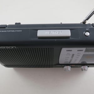 May include: A dark gray Emerson AM/FM/Weather Band portable radio. The radio has a silver "WEATHER" button, a yellow "LIGHT" button, and a telescoping antenna. The radio has a digital clock display and tuning knobs.