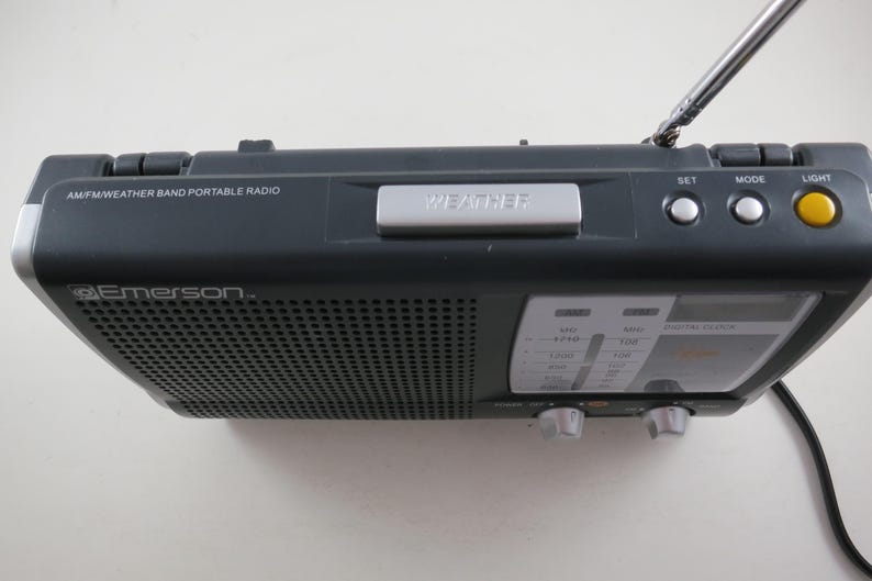 May include: A dark gray Emerson AM/FM/Weather Band portable radio. The radio has a silver "WEATHER" button, a yellow "LIGHT" button, and a telescoping antenna. The radio has a digital clock display and tuning knobs.