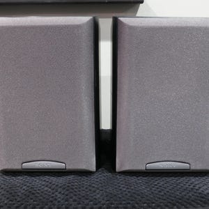 May include: Two gray Sony speakers with a textured finish. Each speaker has a rectangular shape with rounded edges and a small, silver-colored panel at the bottom with the Sony logo. The speakers are placed on a dark, textured surface.