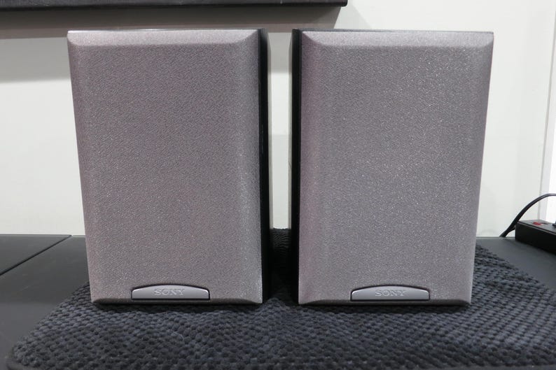 May include: Two gray Sony speakers with a textured finish. Each speaker has a rectangular shape with rounded edges and a small, silver-colored panel at the bottom with the Sony logo. The speakers are placed on a dark, textured surface.