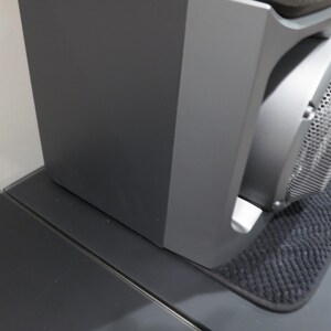 May include: A dark gray audio speaker system with a mesh front. The speaker is placed on a dark gray surface, with a small black mat underneath. The speaker is next to a white wall.