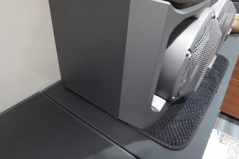 May include: A dark gray audio speaker system with a mesh front. The speaker is placed on a dark gray surface, with a small black mat underneath. The speaker is next to a white wall.