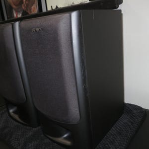 May include: A pair of black Sony bookshelf speakers with rectangular fabric grilles covering the drivers and a bass reflex port located at the bottom front of each unit. The speakers feature a matte black finish and a vertical orientation.