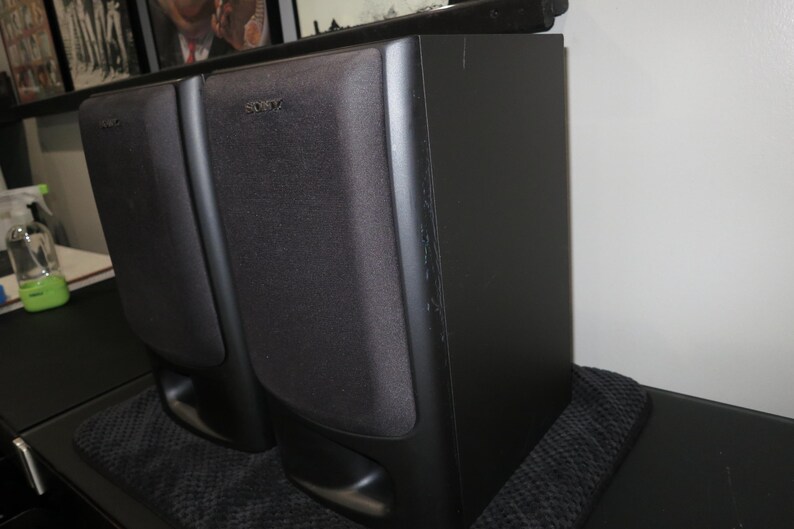 May include: A pair of black Sony bookshelf speakers with rectangular fabric grilles covering the drivers and a bass reflex port located at the bottom front of each unit. The speakers feature a matte black finish and a vertical orientation.