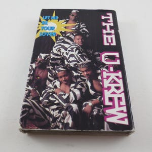 May include: A vintage cassette tape featuring the group "The U-Krew." The cover art displays a group of people in black and white striped outfits. The text "LET ME BE YOUR LOVER" and "THE U-KREW" are also visible.