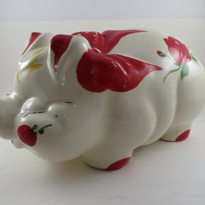 May include: A ceramic piggy bank in the shape of a pig, painted white with red accents. The pig has red spots on its back and red feet. It also features hand-painted red roses and green leaves.