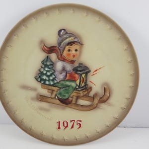 May include: A vintage 1975 Christmas plate featuring a child on a sled with a lantern. The plate is decorated with a cream background and a brown border with a star pattern. The child is wearing a blue hat, a purple jacket, and a red scarf. The sled is brown and has a green tree on it.