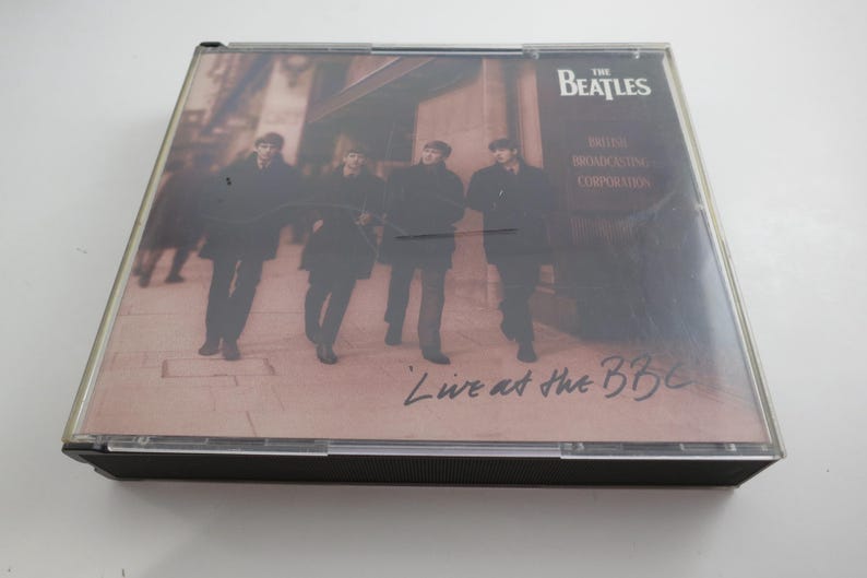 May include: A CD case featuring The Beatles' album "Live at the BBC." The cover art shows the band members walking on a street. The album title and band name are visible on the cover.